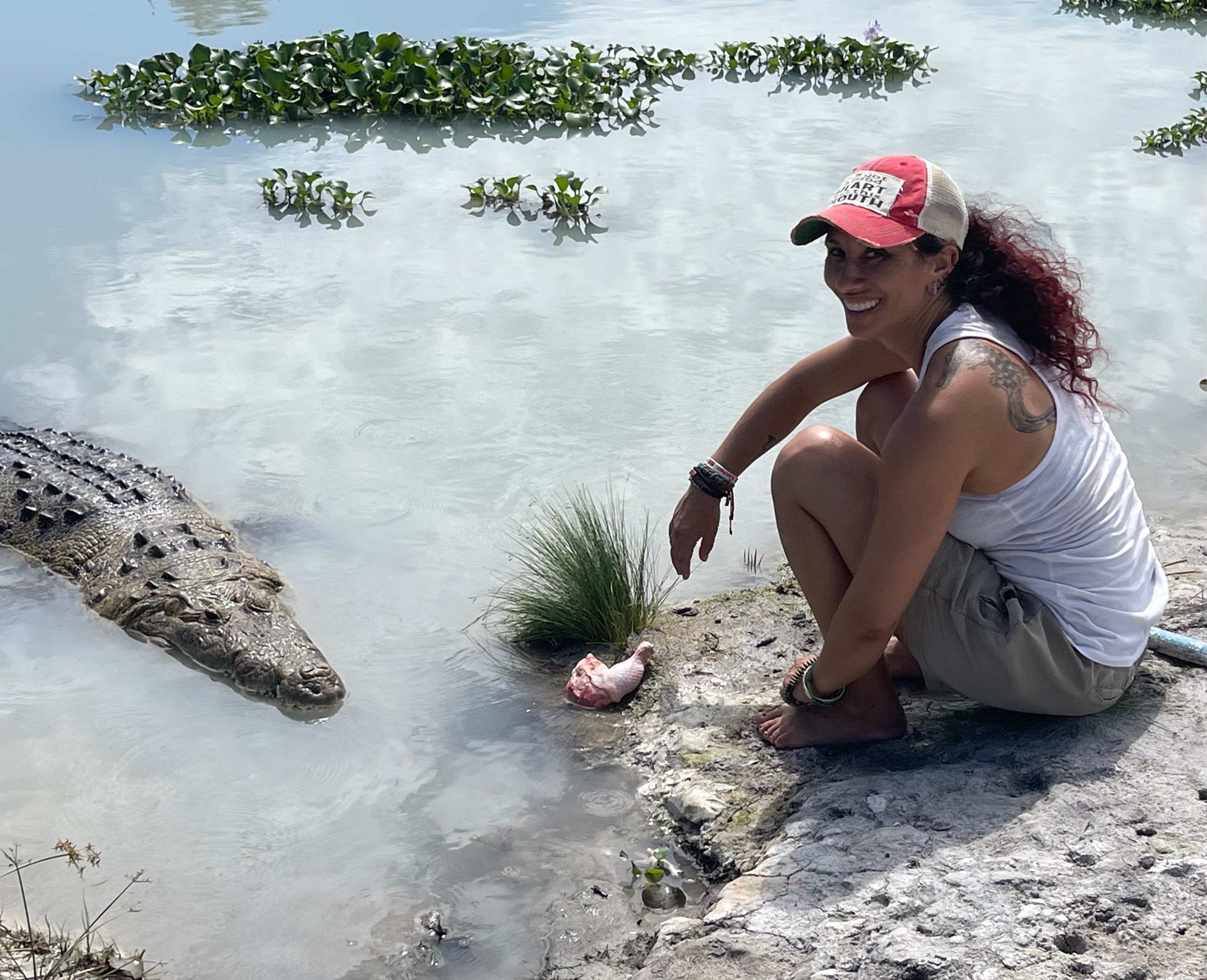 A woman sits ont eh shore of a body of water, where a croc sits about 5 feet away. A chicken leg sits at her feet.