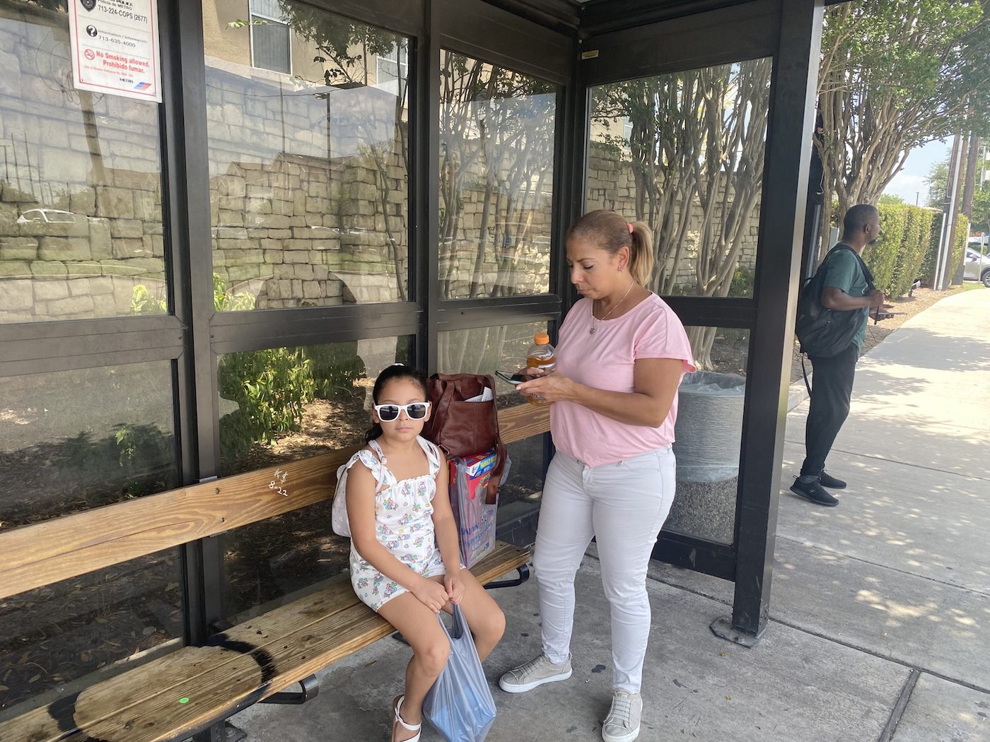 A woman standing underneath a bus shelter. Her young daughter wears sunglasses, sitting on the bench. 