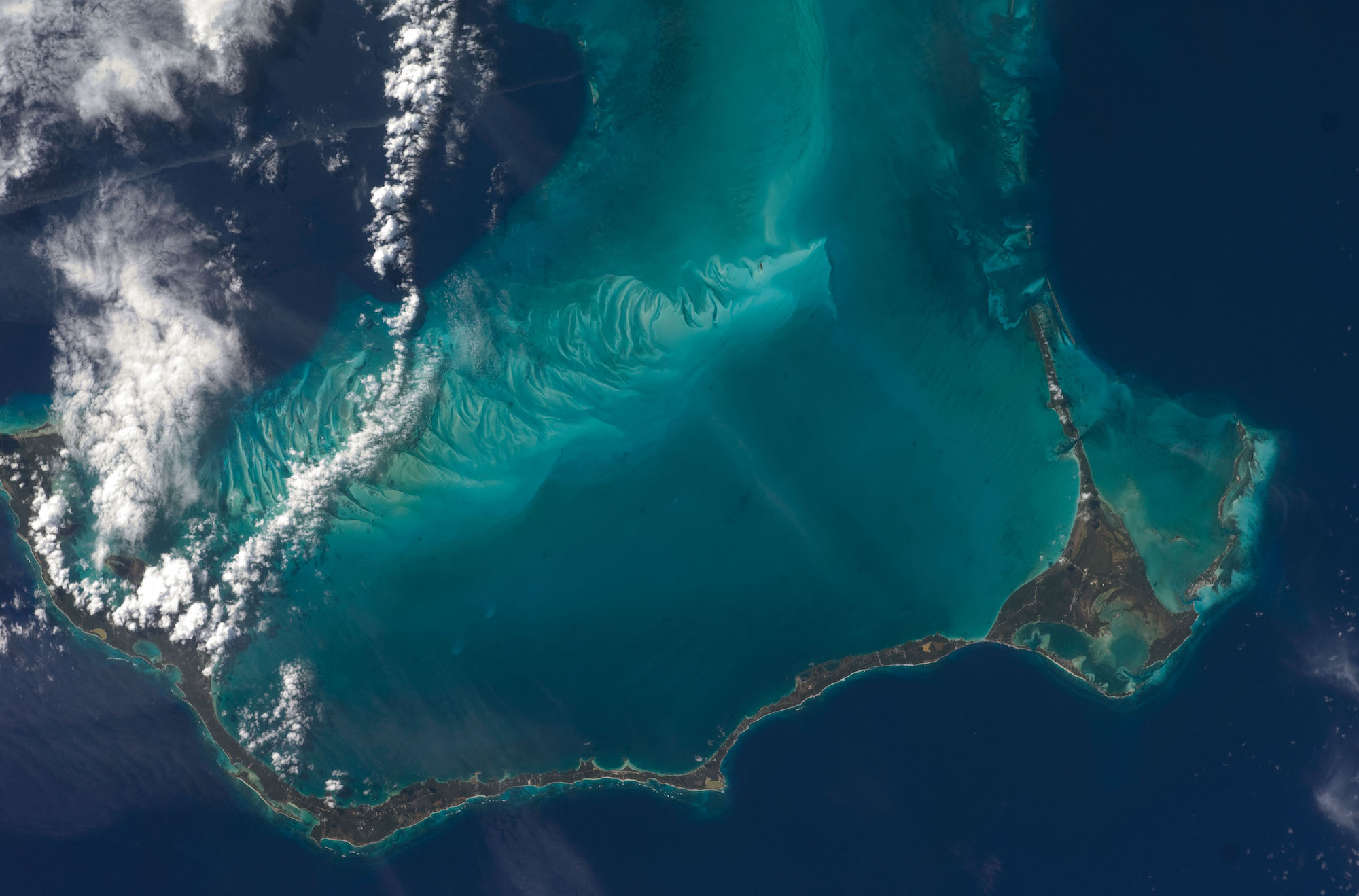 Aerial view of a long, thin, curved island amid dark blue and turquoise waters, a few white clouds down the left side