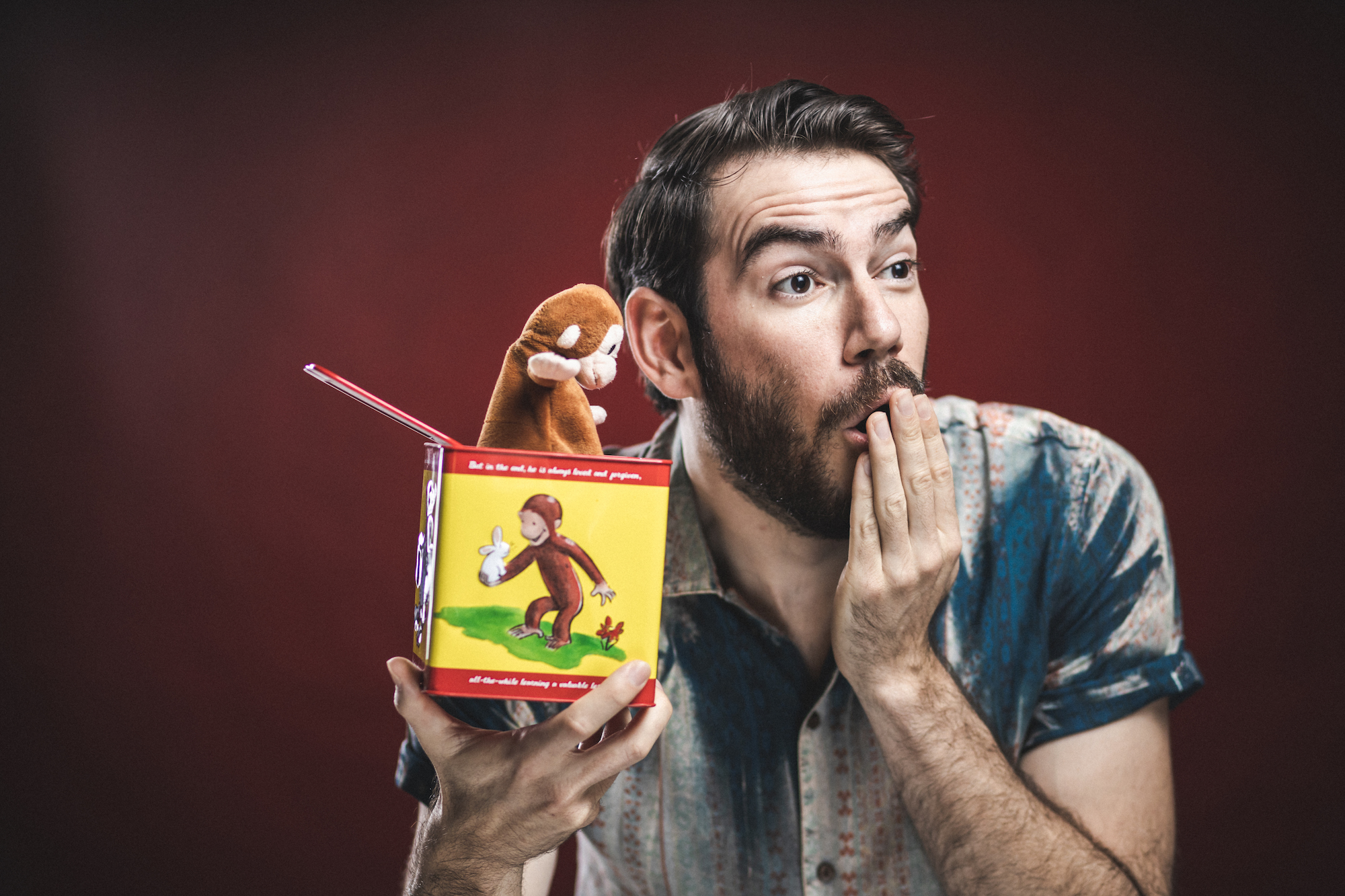 Man with light skin holds a Curious George jack-in-the-box and covers his mouth with one hand, surprised