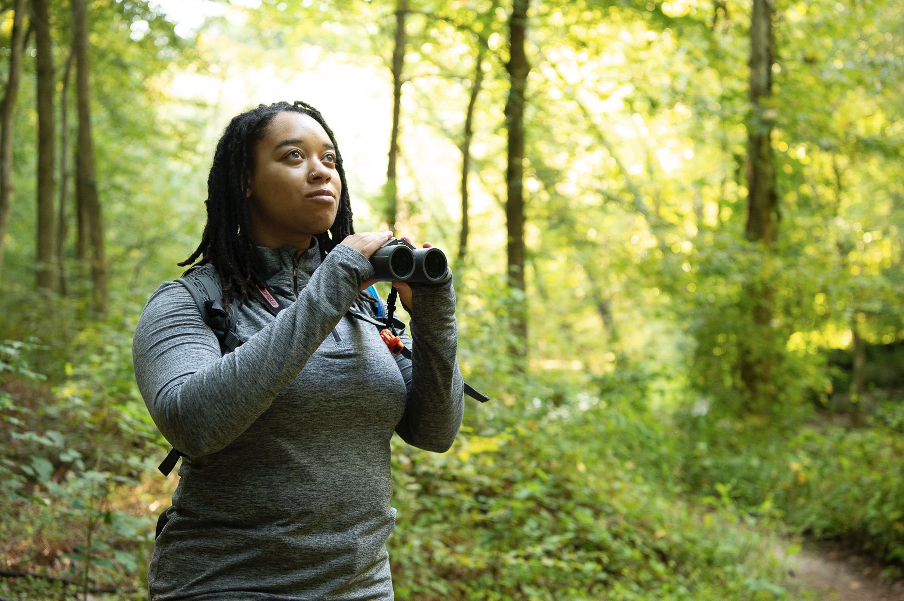 A woman holds up binoculars in a green forest.
