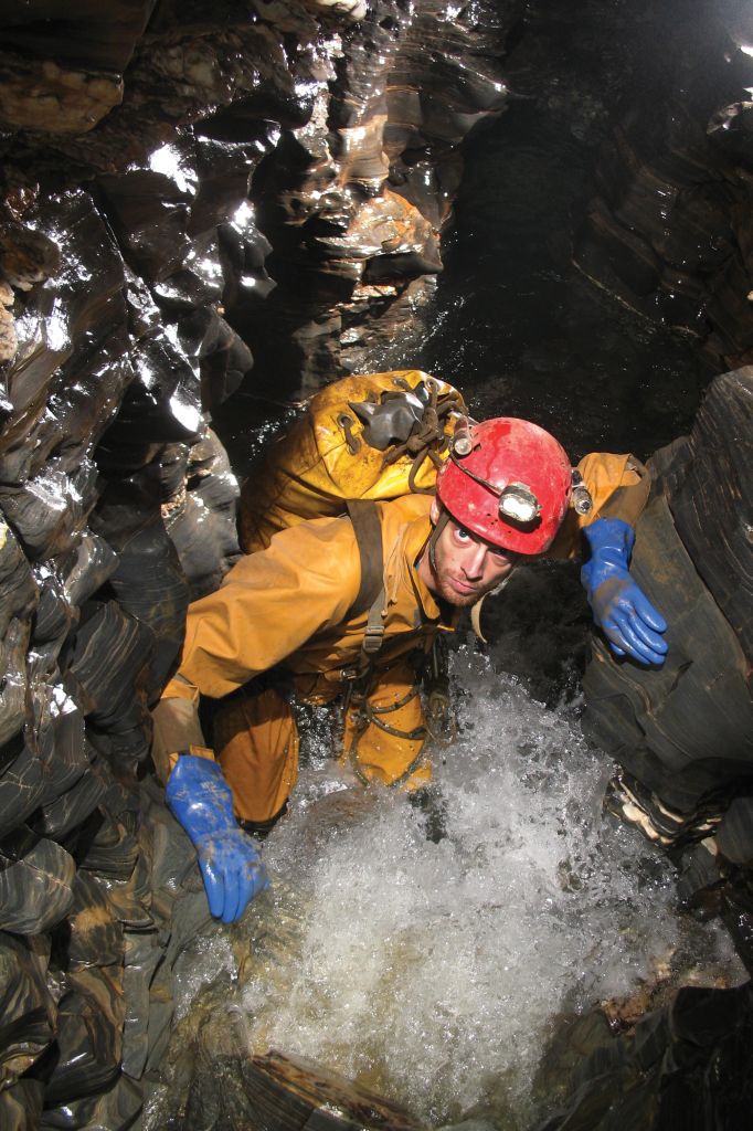 A man wearing a yellow jumpsuit and a helmet stands inside a cave full of water.