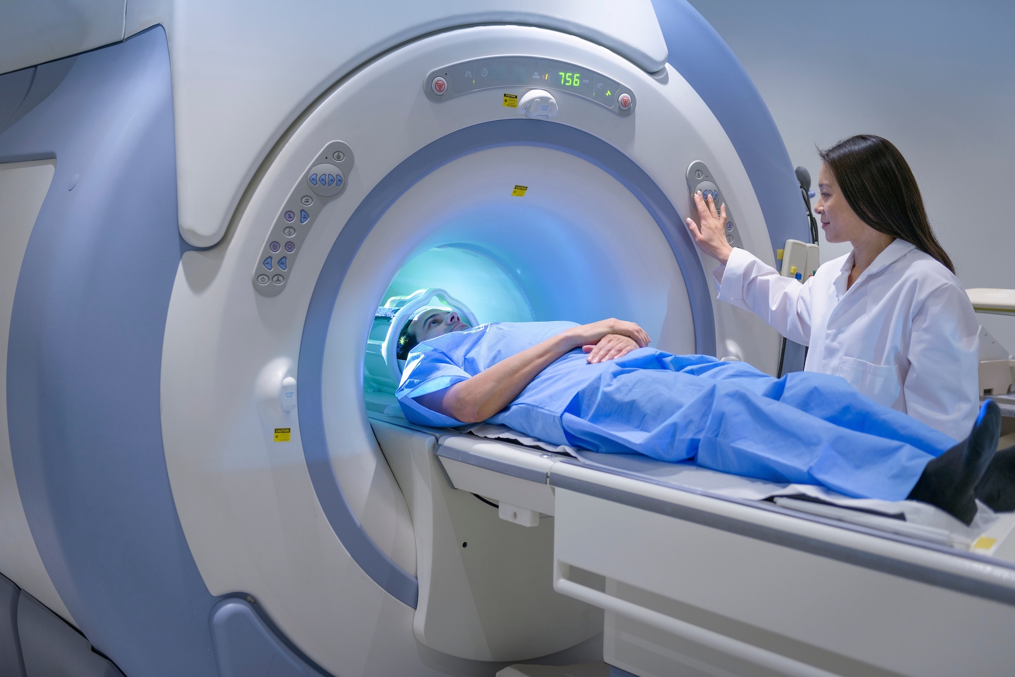 A male patient in blue scrubs lies on a table with his head inside the large cynlinder of a blue and white MRI machine. A female doctor in a white coat stands next to him in with her right hand on the machine's buttons.