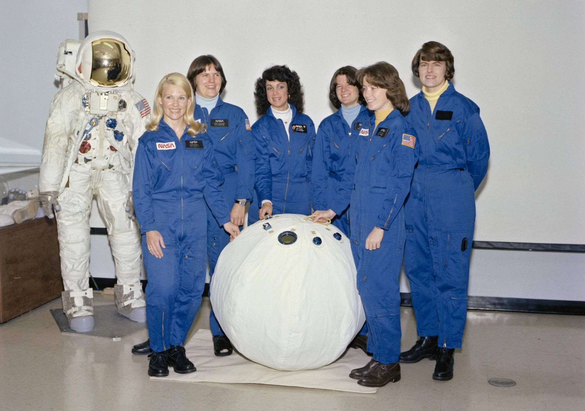 Six women stand in front of a white ball, with a mannequin wearing a space suit among them.