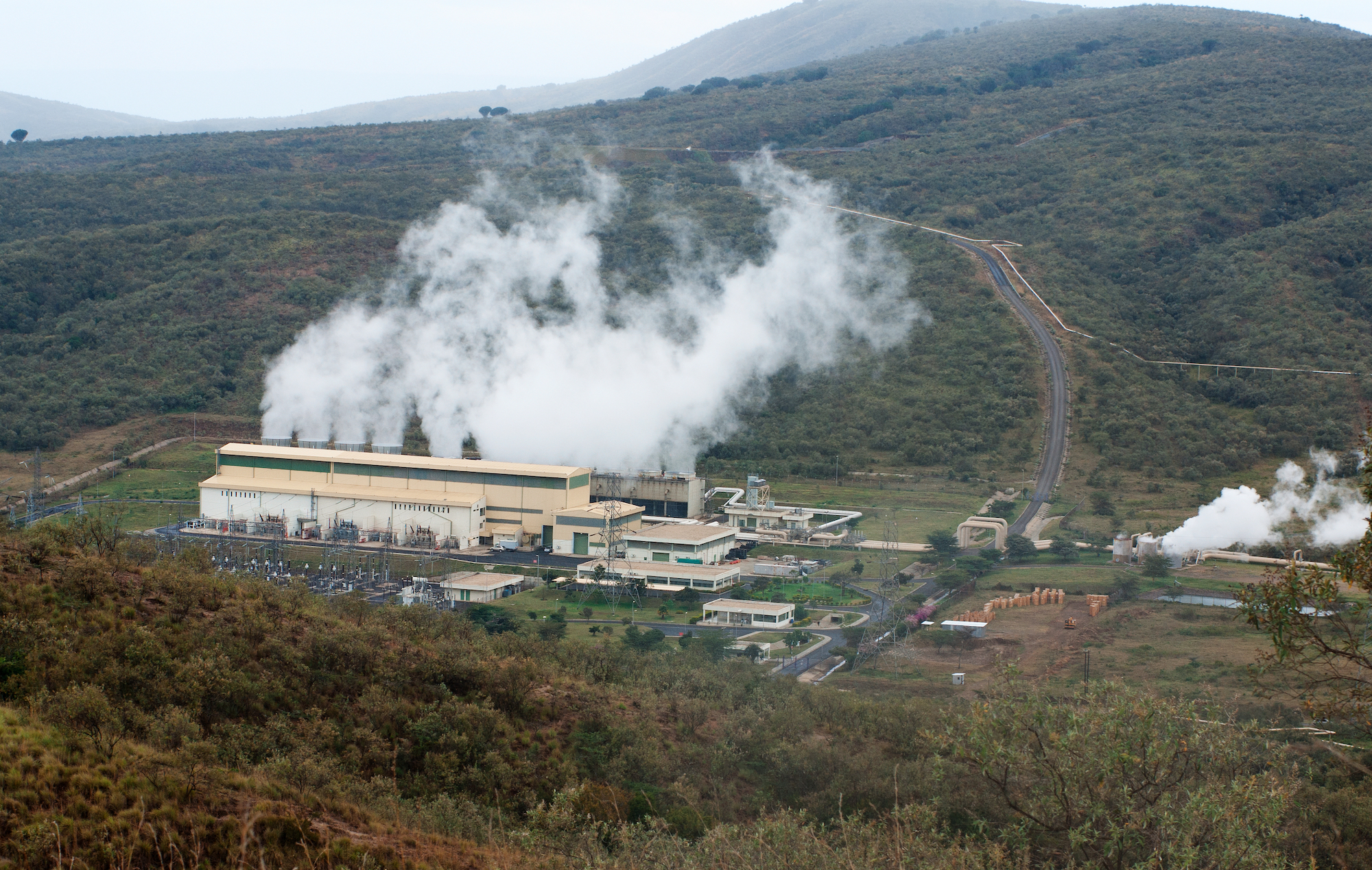Plumes of steam drift up from a line of cooling towers behind a long, beige building. The structures are set in green, arid valley between two hills.