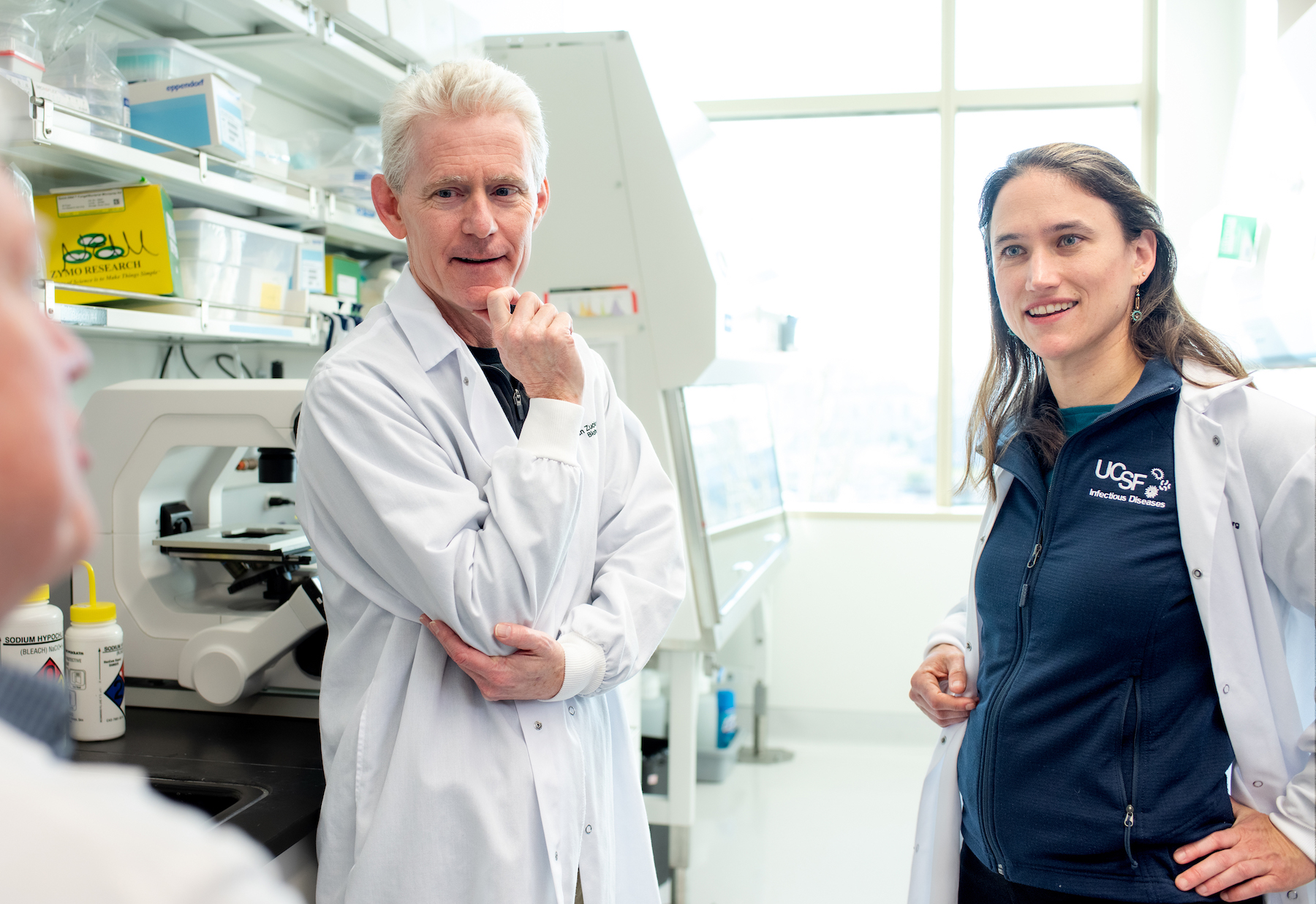 Two doctors stand in a lab, wearing lab coats. Joe DeRisi is on the left, and Natasha Spottiswoode is on the right.