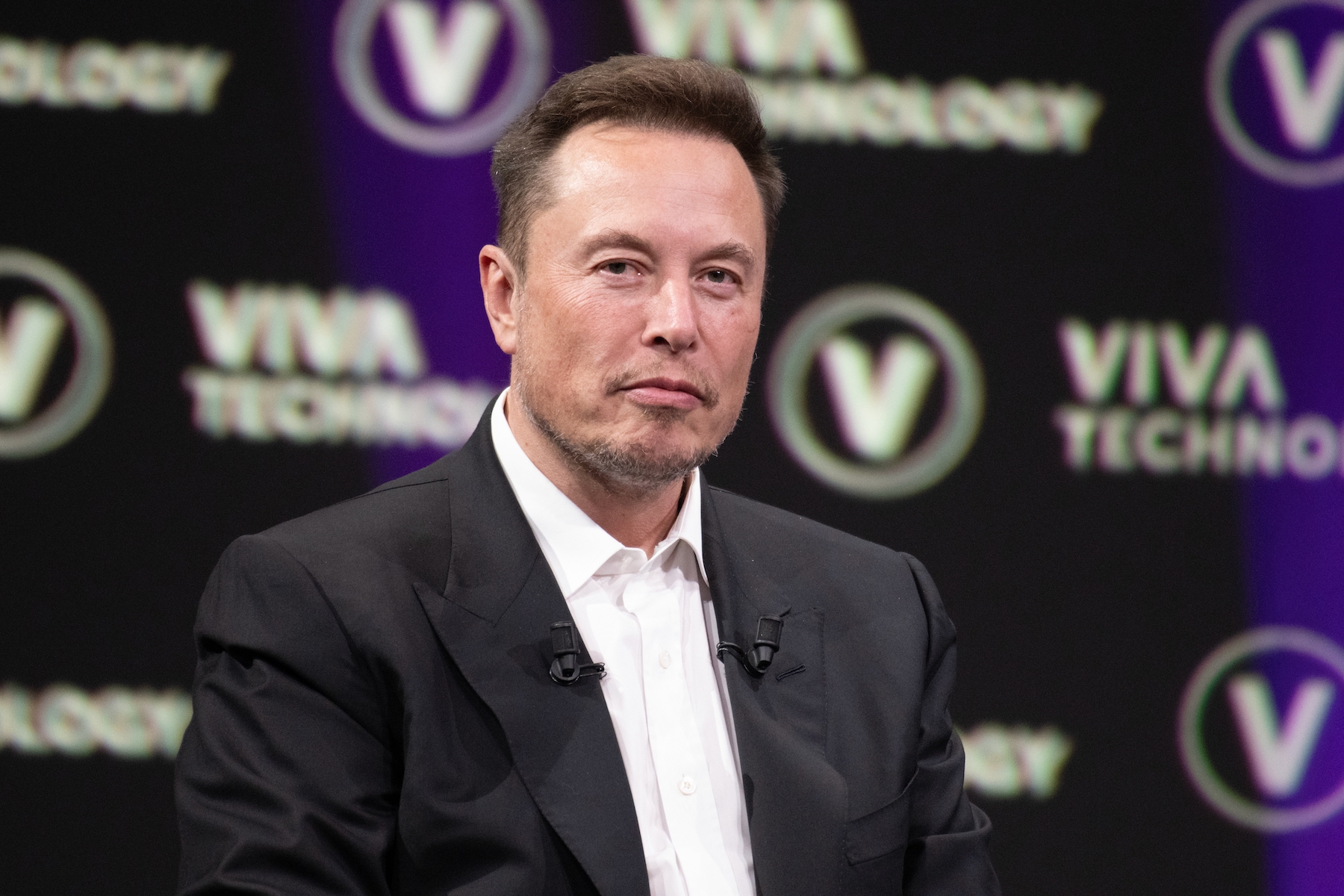 Elon Musk, looking into the camera sitting at a press conference. Bust shot.