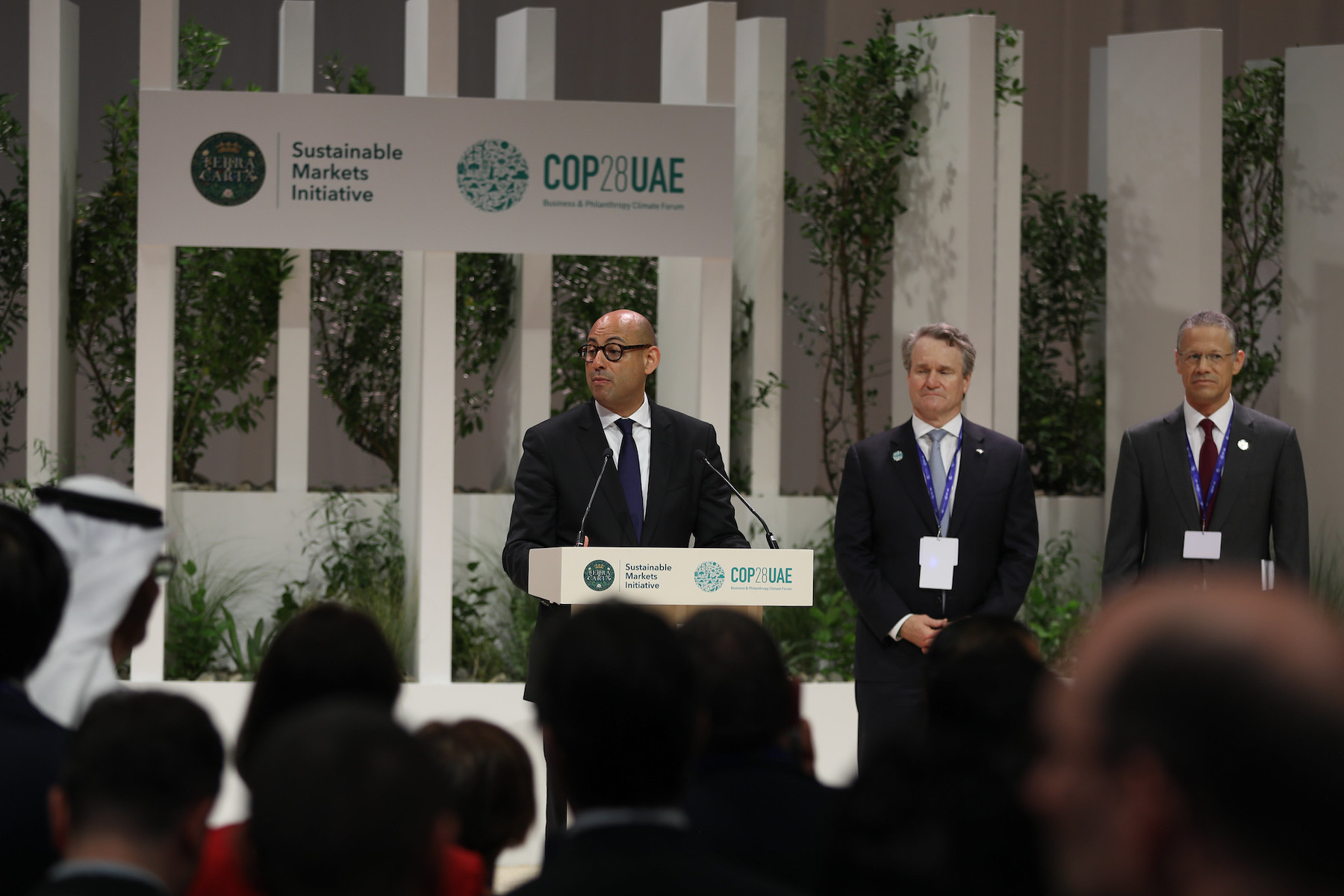 A man stands at a podium speaking to a crowd. Behind him is a sign that says "COP28UAE"