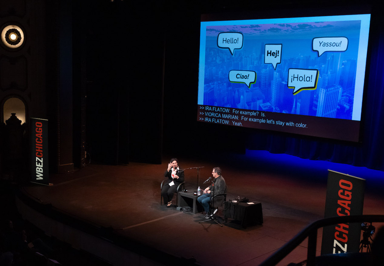 Ira Flatow and scientist Viorica Marian sit on a stage. Behind them is a screen with speech bubbles containing the word "hello" in multiple languages.