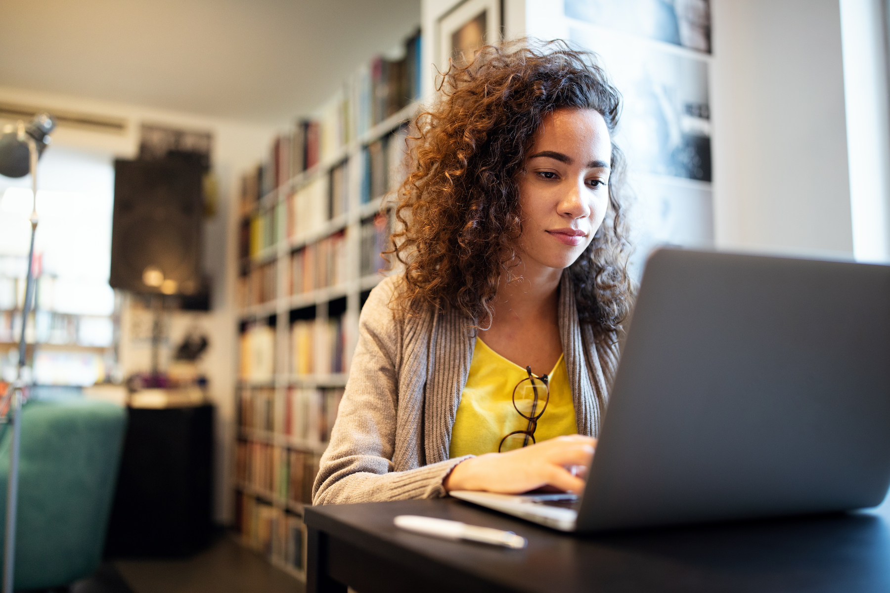 A young woman with dark curly hair wearing a yellow shirt sits in a library typing on a laptop