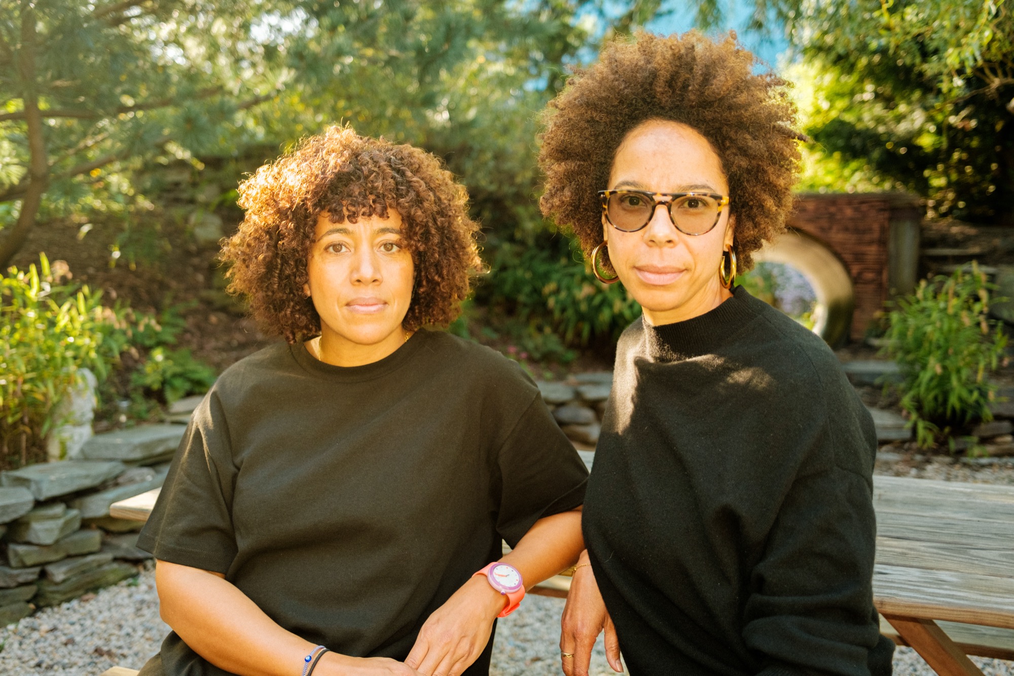 Two Black women wearing black shirts sit in a garden on a sunny day, looking into the camera