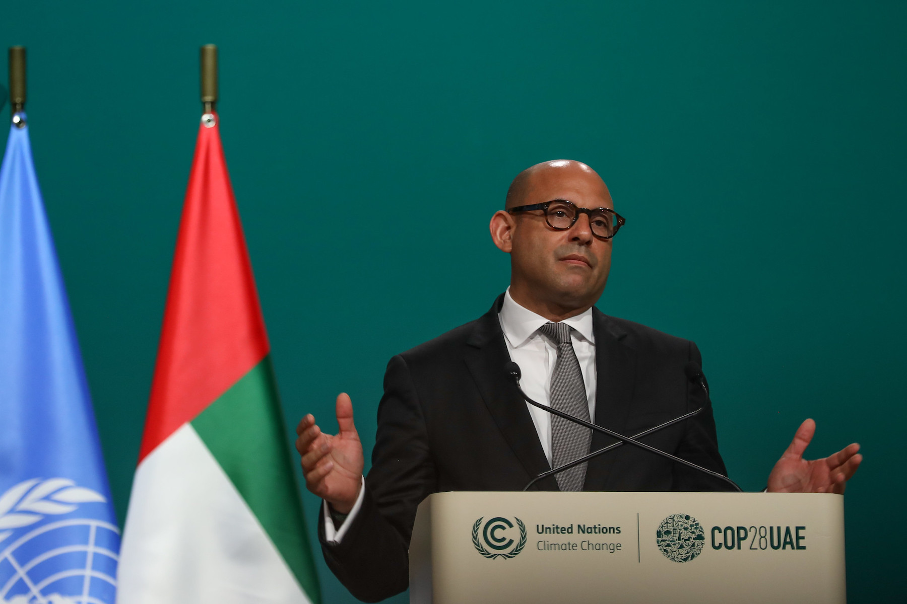 A man in a suit gives a speech, looking tired against a green background