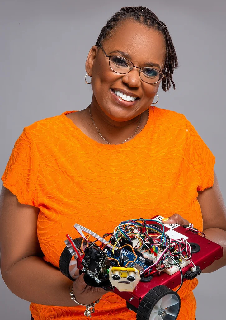 A Black woman smiling at the camera, holding up a robotics project.