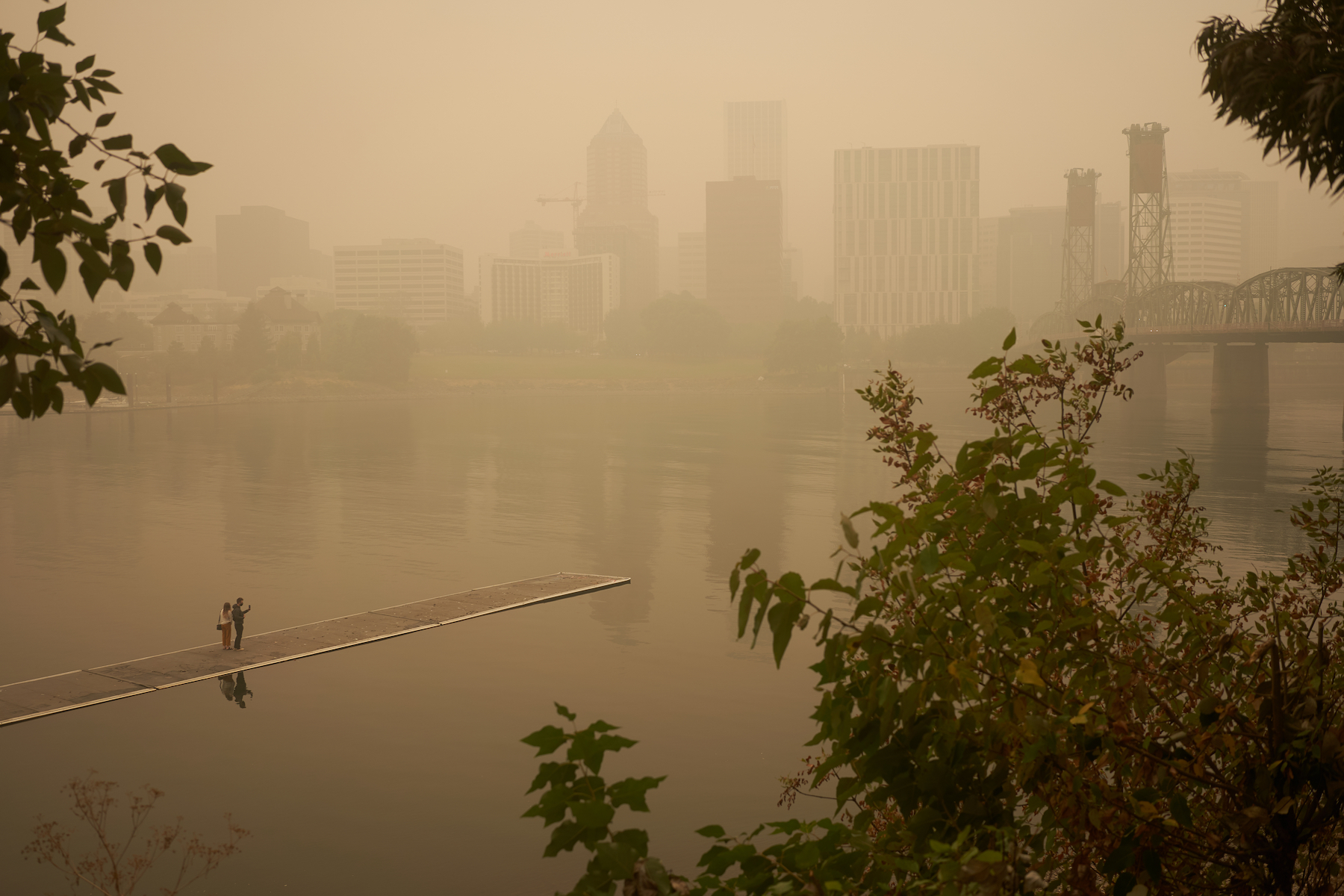 A city skyline barely visible through thick grey smoke. Two people stand on a dock photographing it.