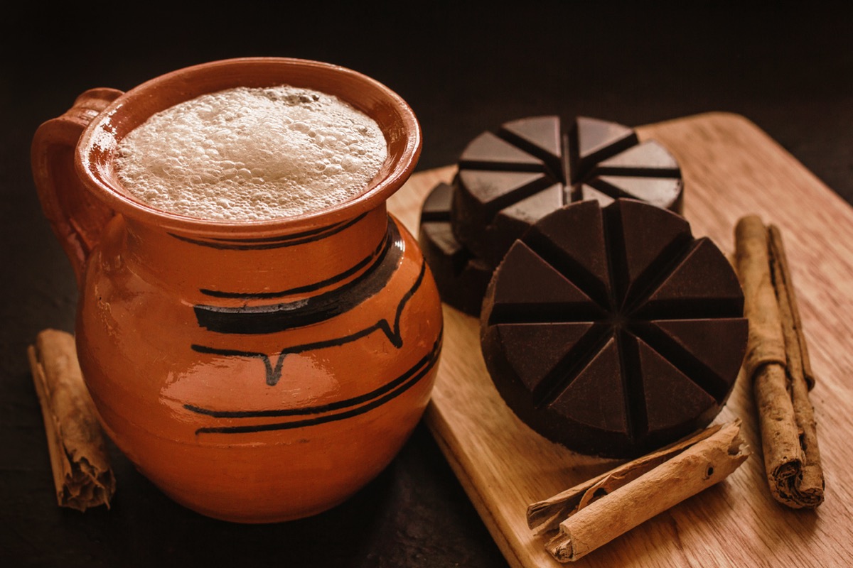 A frothy beverage in a rustic brown mug sits next to cinnamon sticks and dark disks of chocolate.