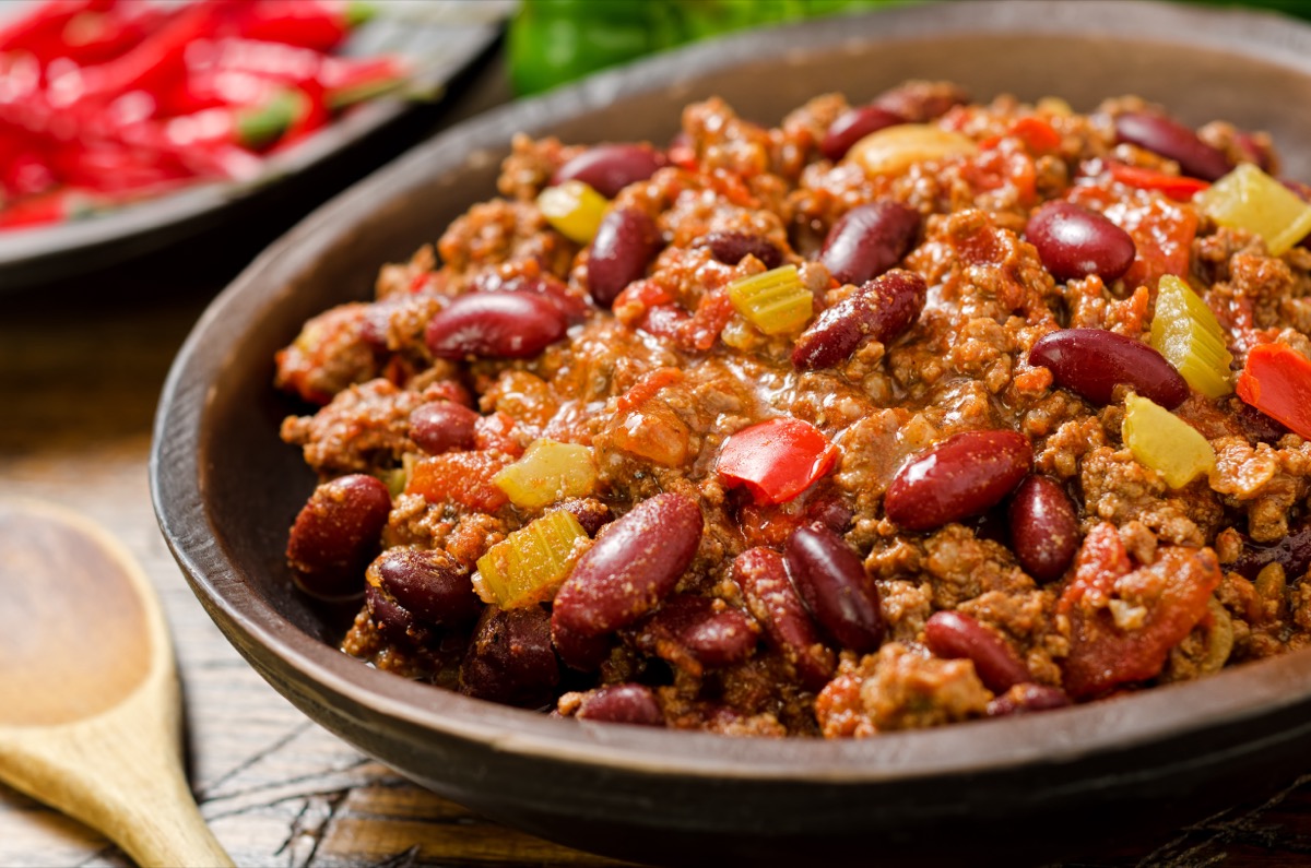 A bowl of chili con carne in a shallow brown bowl on a wooden table with a wooden spoon nearby, and chili peppers in the background.