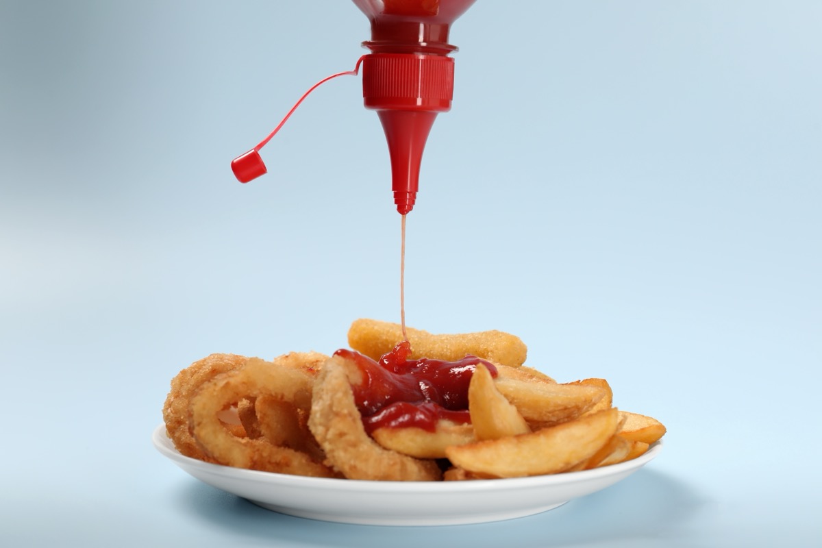 Pouring ketchup on to french fries and fried onion rings against a light blue background.