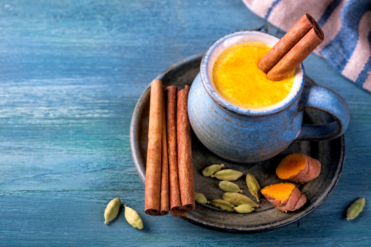 A creamy, frothy golden-colored beverage in a blue ceramic mug sits on a plate that holds fresh turmeric, cinnamon sticks, and cardamom pods.