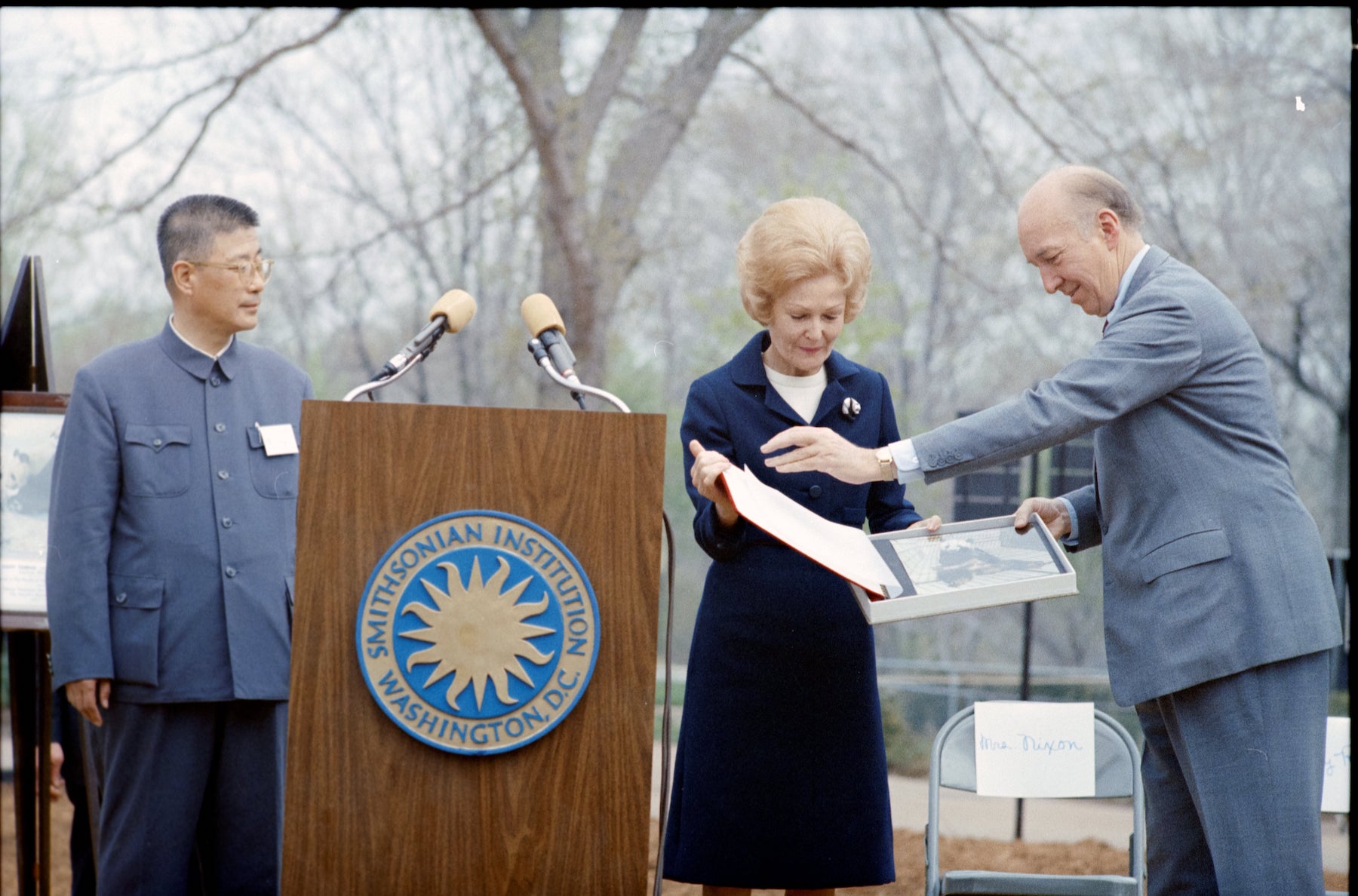 A white man and woman open a photo album of pandas by a podium with the Smithsonian's logo on it. A Chinese man stands behind the podium, looking at the others.