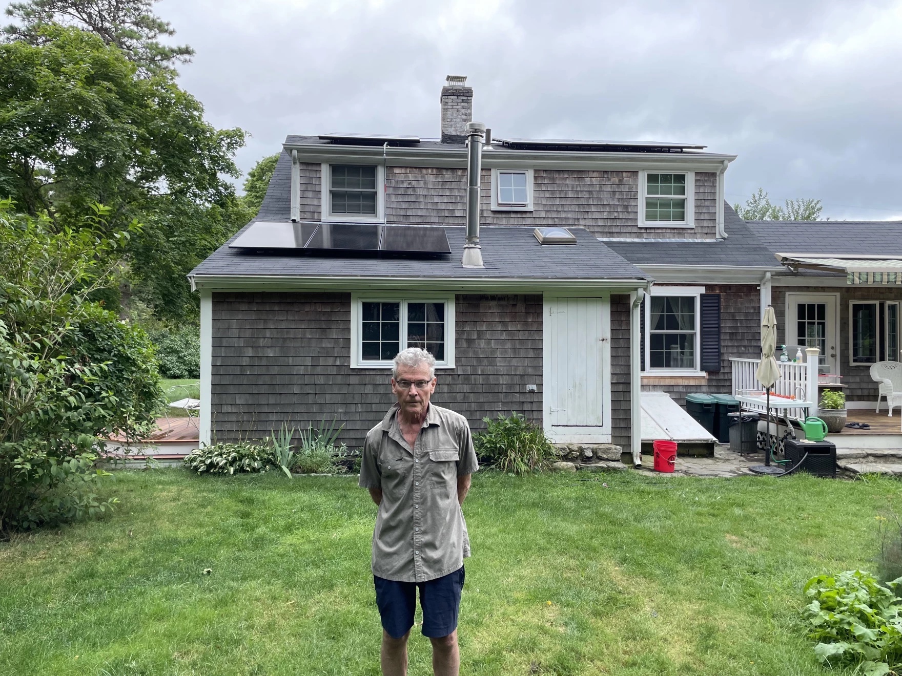 A man stands on the lawn in front of a house with 3 black solar panels on the roof.