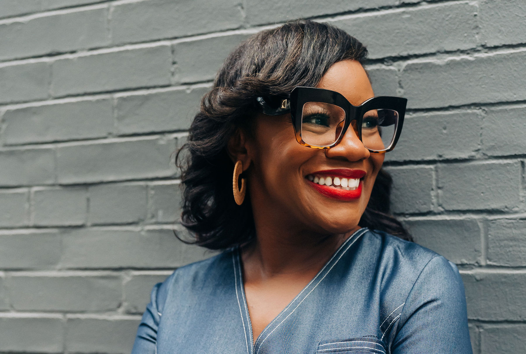 A Black woman wearing glasses smiles in front of a neutral brick background.