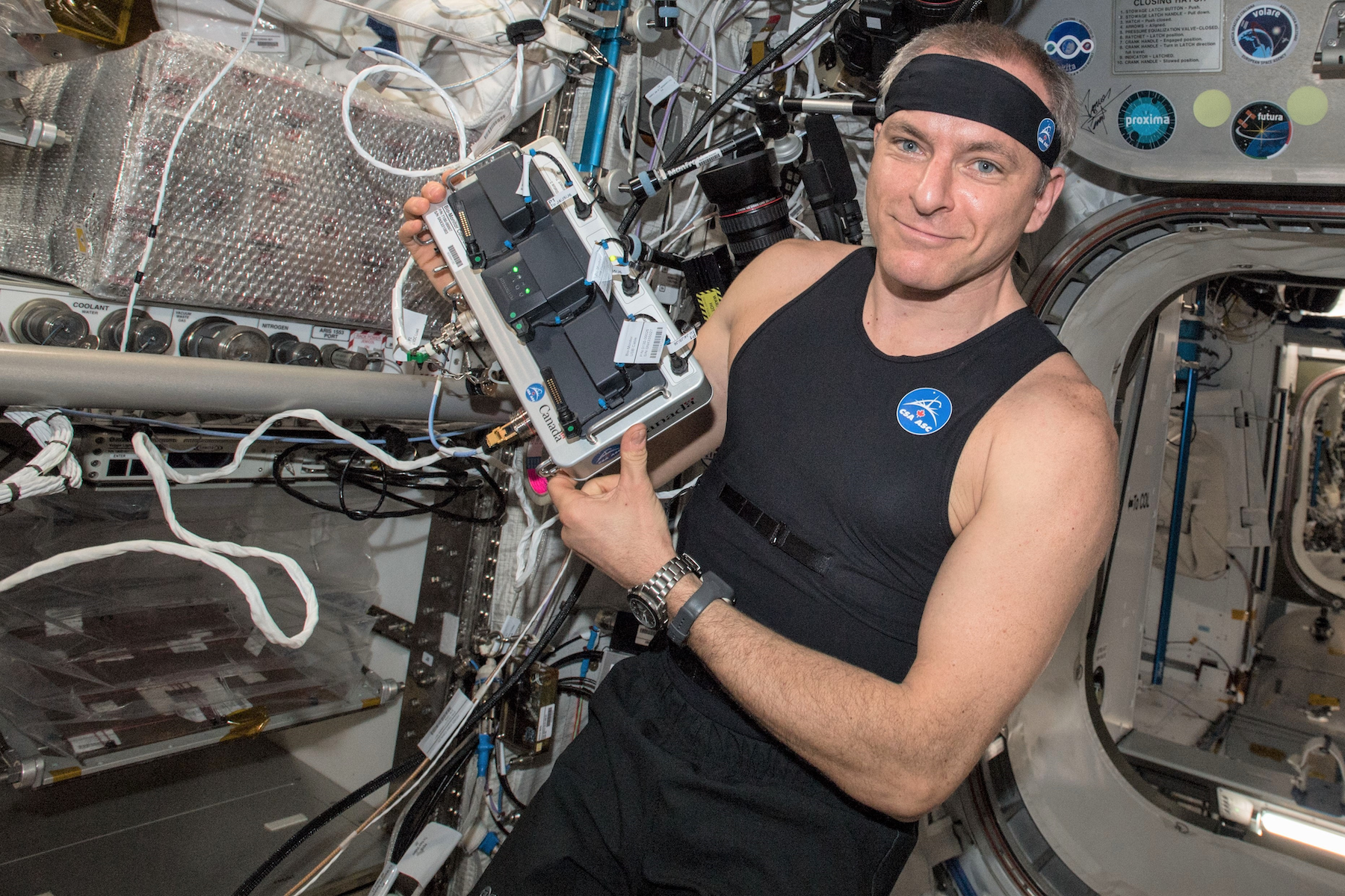 A man wearing a headband in a spaceship holds a complicated-looking rectangular machine.