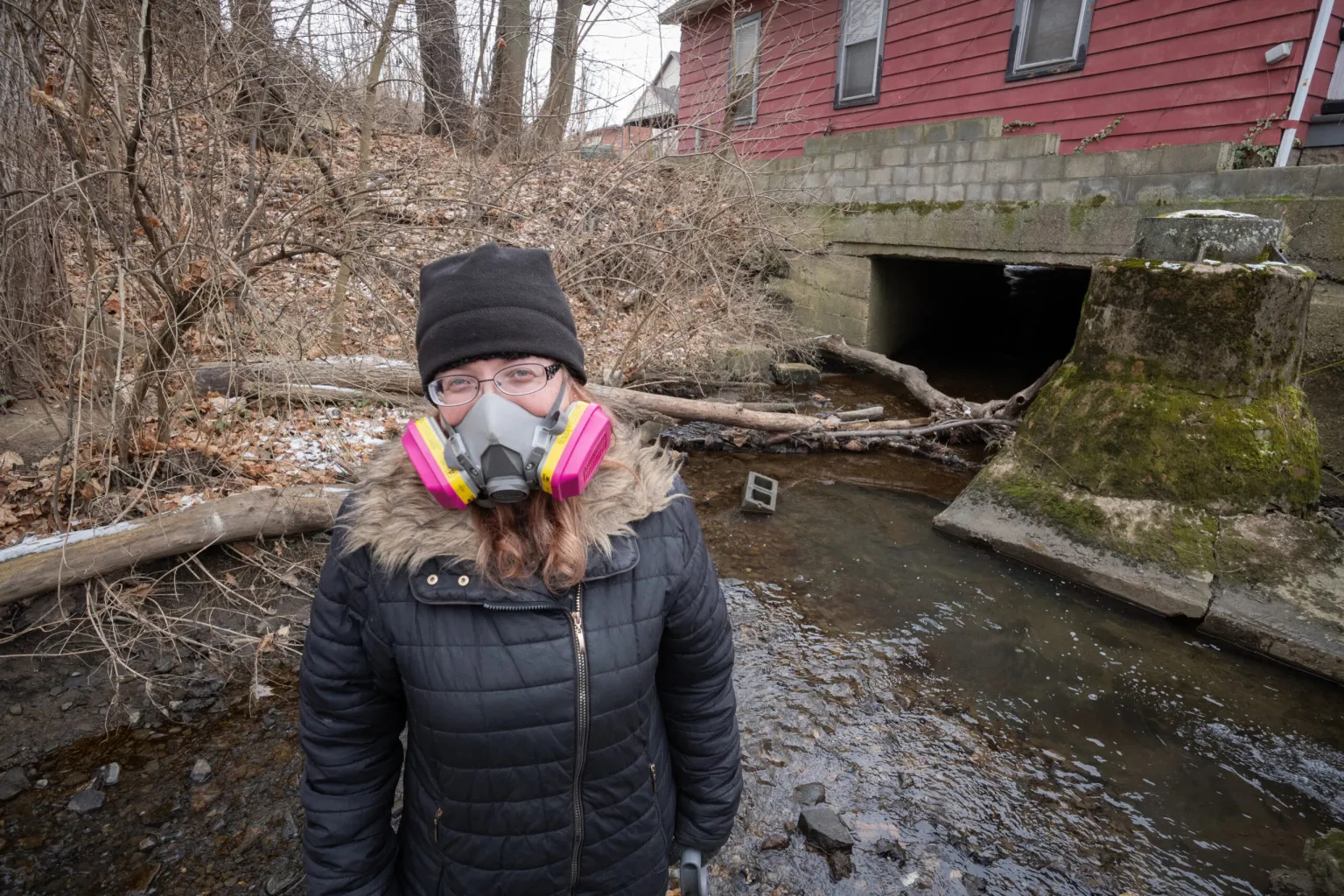 A woman wearing a gas mask over her mouth and nose standing in front of a muddy, polluted body of water.