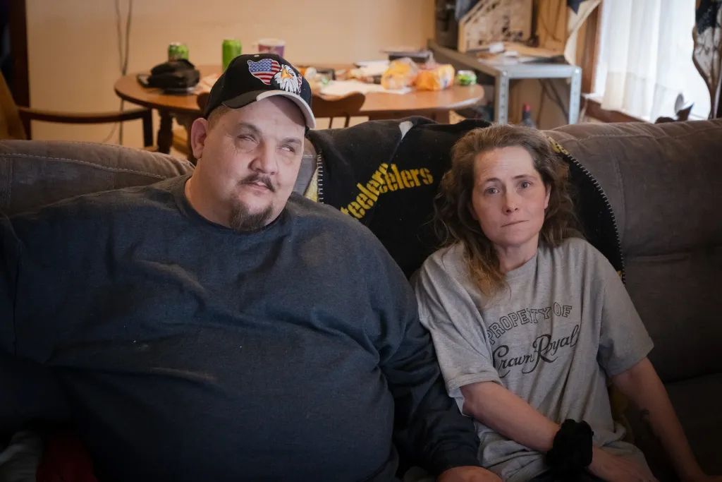 A man sitting next to a woman looking at the camera. Both are indoors, on a couch.