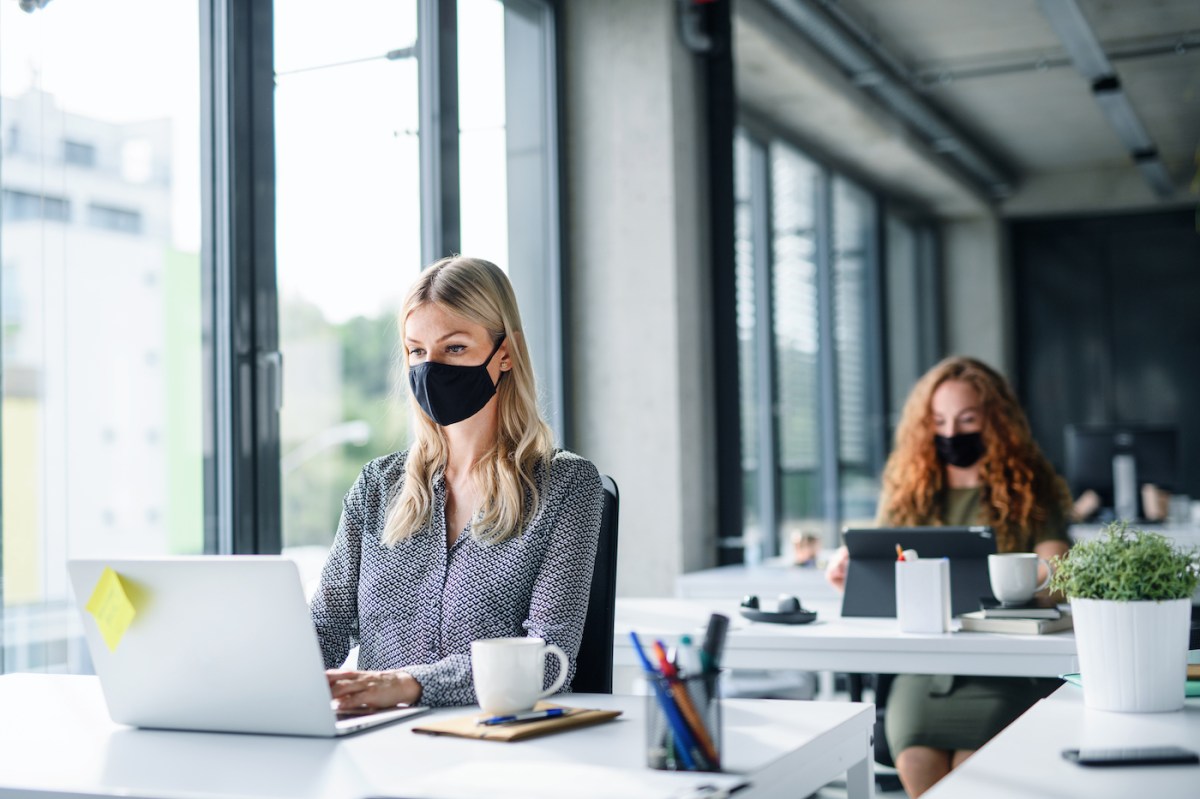 Young people with face masks work at laptops on desks along a wall of windows.