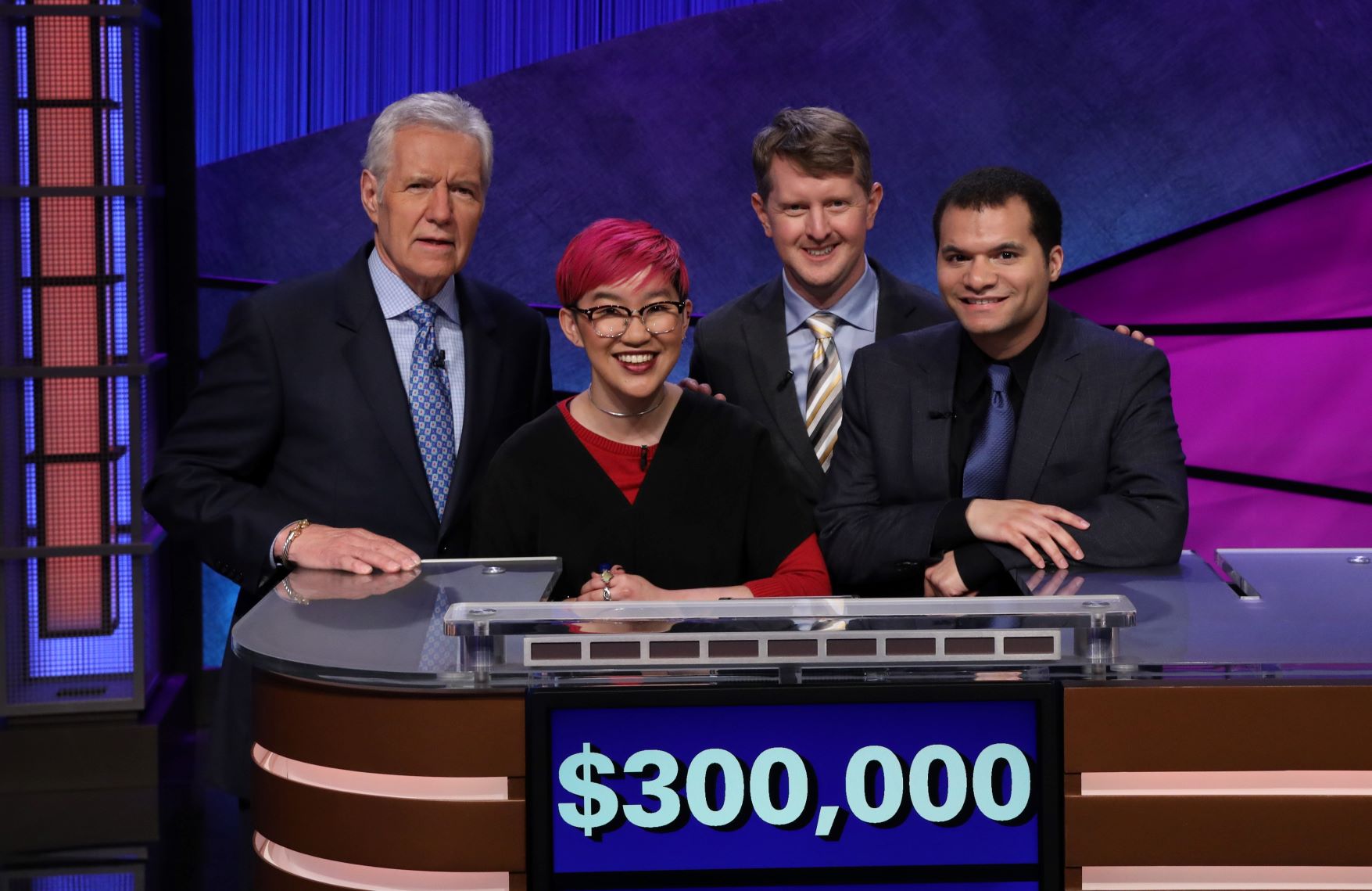Four smiling people standing at a Jeopardy! games desk with "$300,000" on the screen.