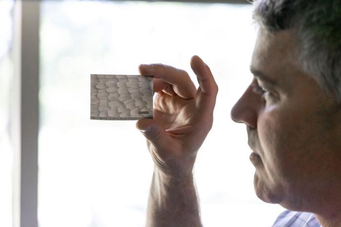 A man holding a small rectangular pane up to a window with a detailed texture etched into it. The texture resembles small patches of horizontal lines, each forming a group that overlaps over another group of lines.