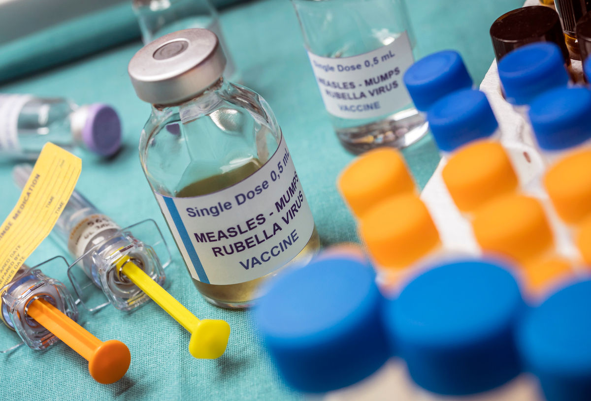 Close up of a medical tray with glass vials of MMR vaccine and syringes lying next to them, on a green fabric.