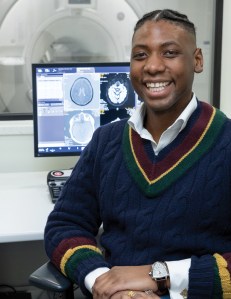 A Black man smiles at the camera. Behind his is an MRI machine and brain scans on a monitor.