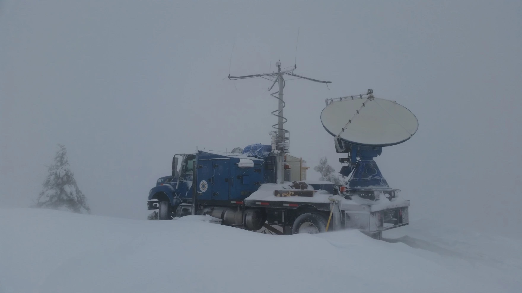 A truck with a large satellite dish in its rear carriage driving through a very snowy area.