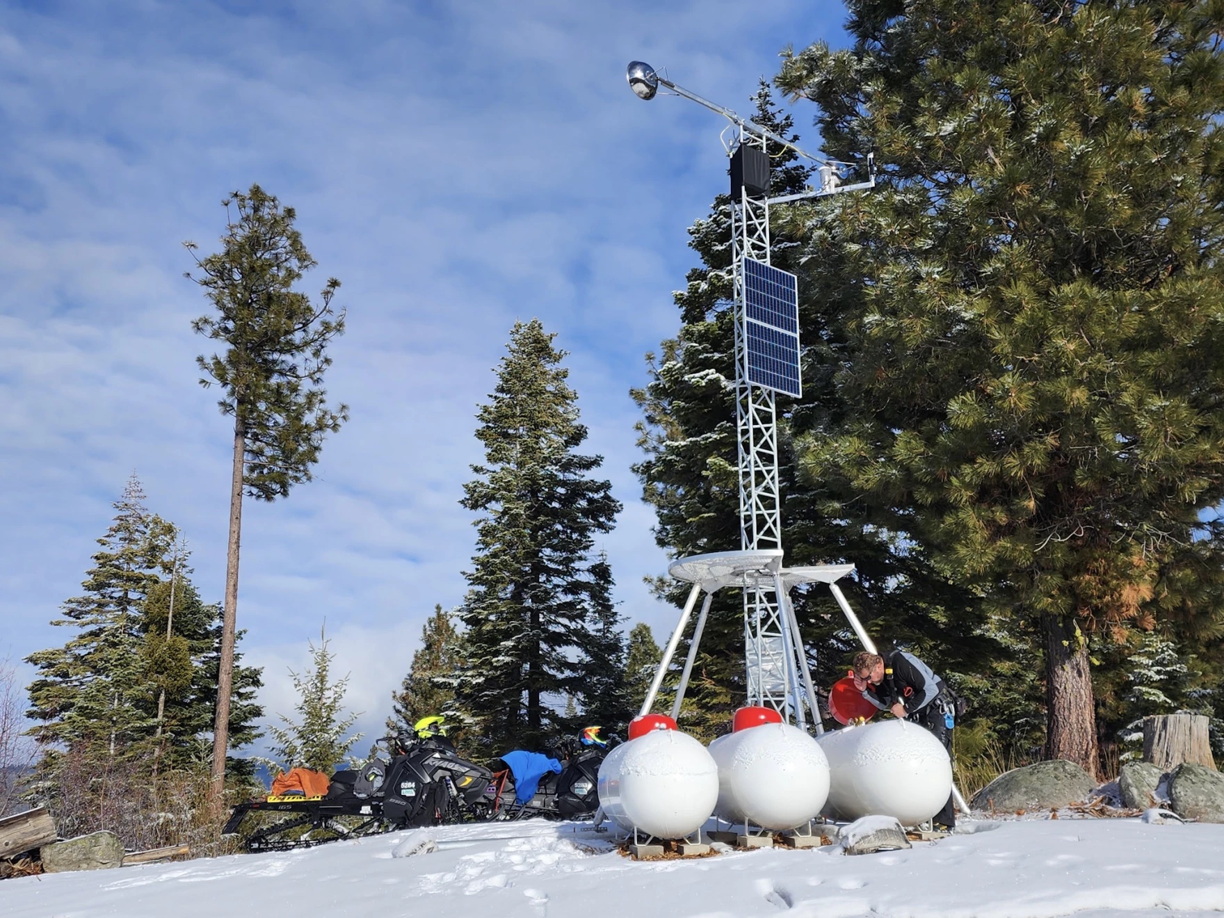 A metal antenna-like object with a solar panel attached to it stands on top of a snowy hill.