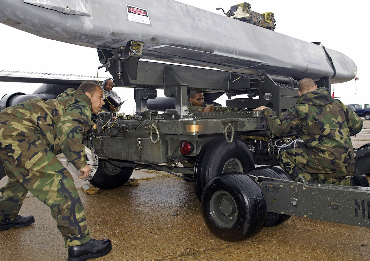 Four men in camo are crouched around the base of a large platform holding a long, gray missile-like object.