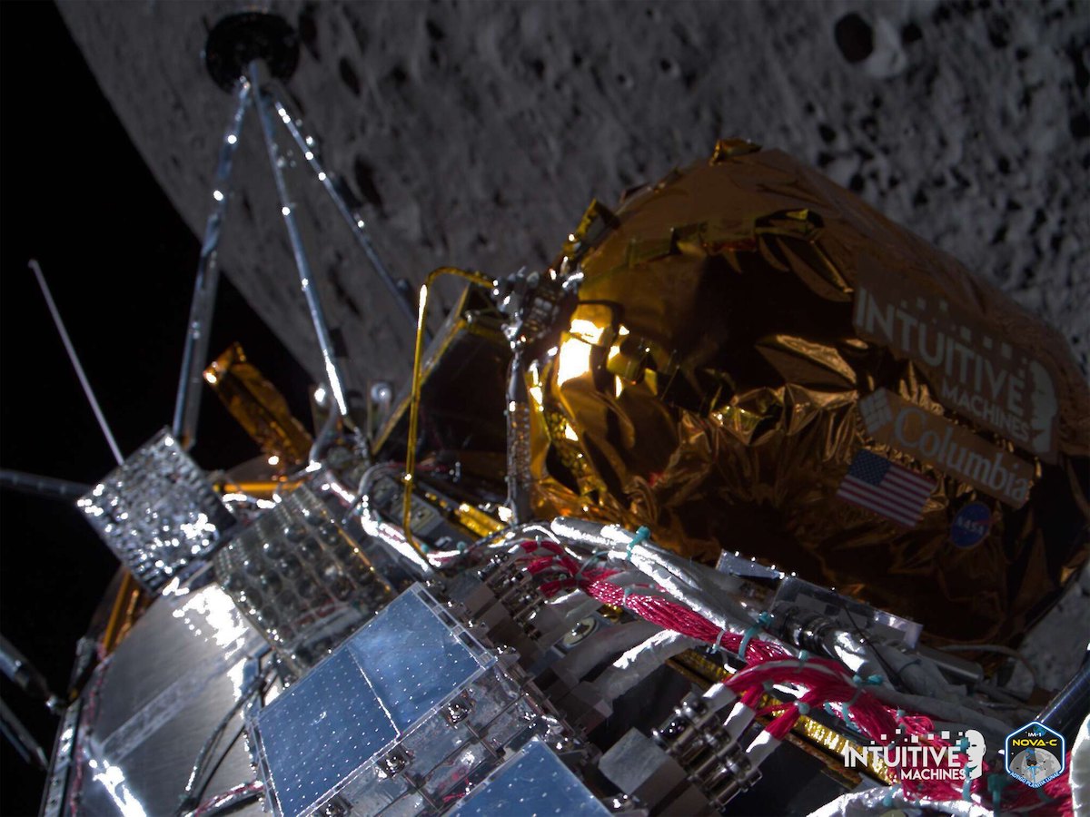 A view down the silvery side of a moon lander with the gray lunar surface very close in the background.