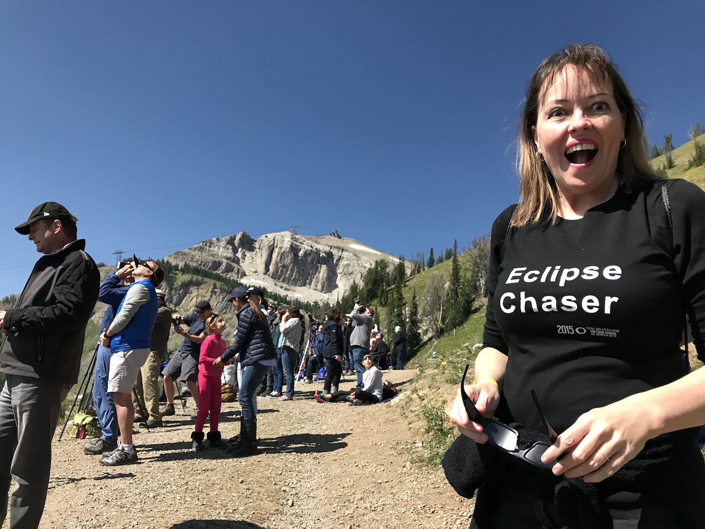 A woman smiles wide-eyed at the camera with a clear sky and many people gathered behind her. She's in a park.