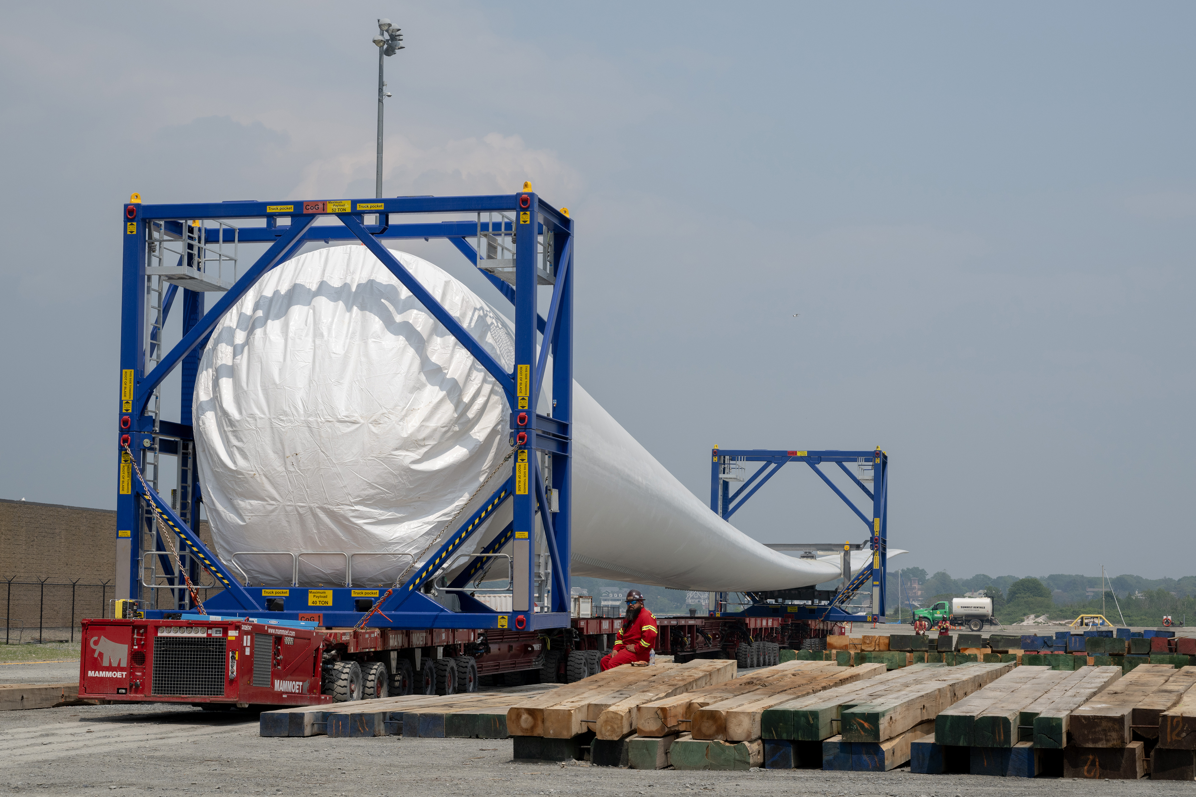 A view down the length of a massive white wind turbine blade held by two large transport devices on the ground as a worker in red looks on.
