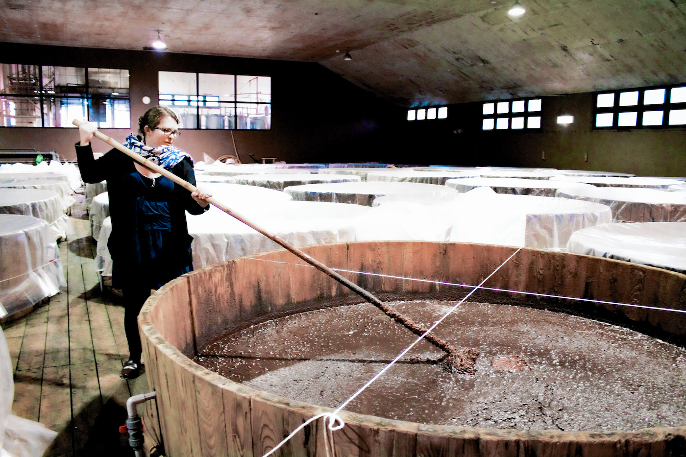 A woman wearing a scarf as she stirs an enormous vat of soy sauce mash.