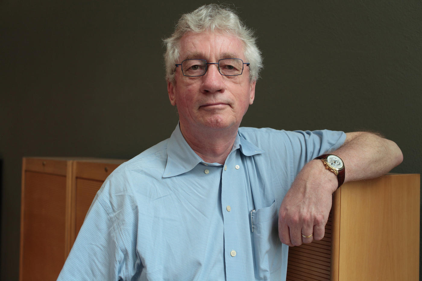 An older man with white hair and glasses looks into the camera. His arm leans on a wooden cabinet.