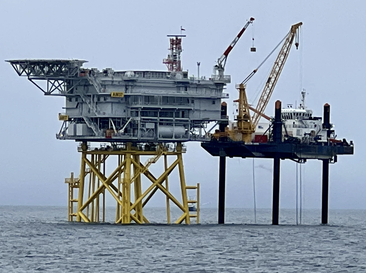 An industrial building stands on yellow stilts above the ocean surface.