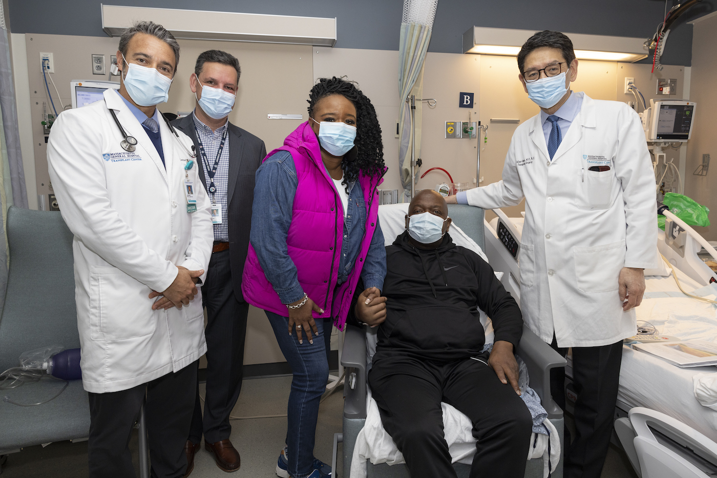 Four people wearing masks stand around a man in a chair, all looking happily at the camera. They're in a hospital room.