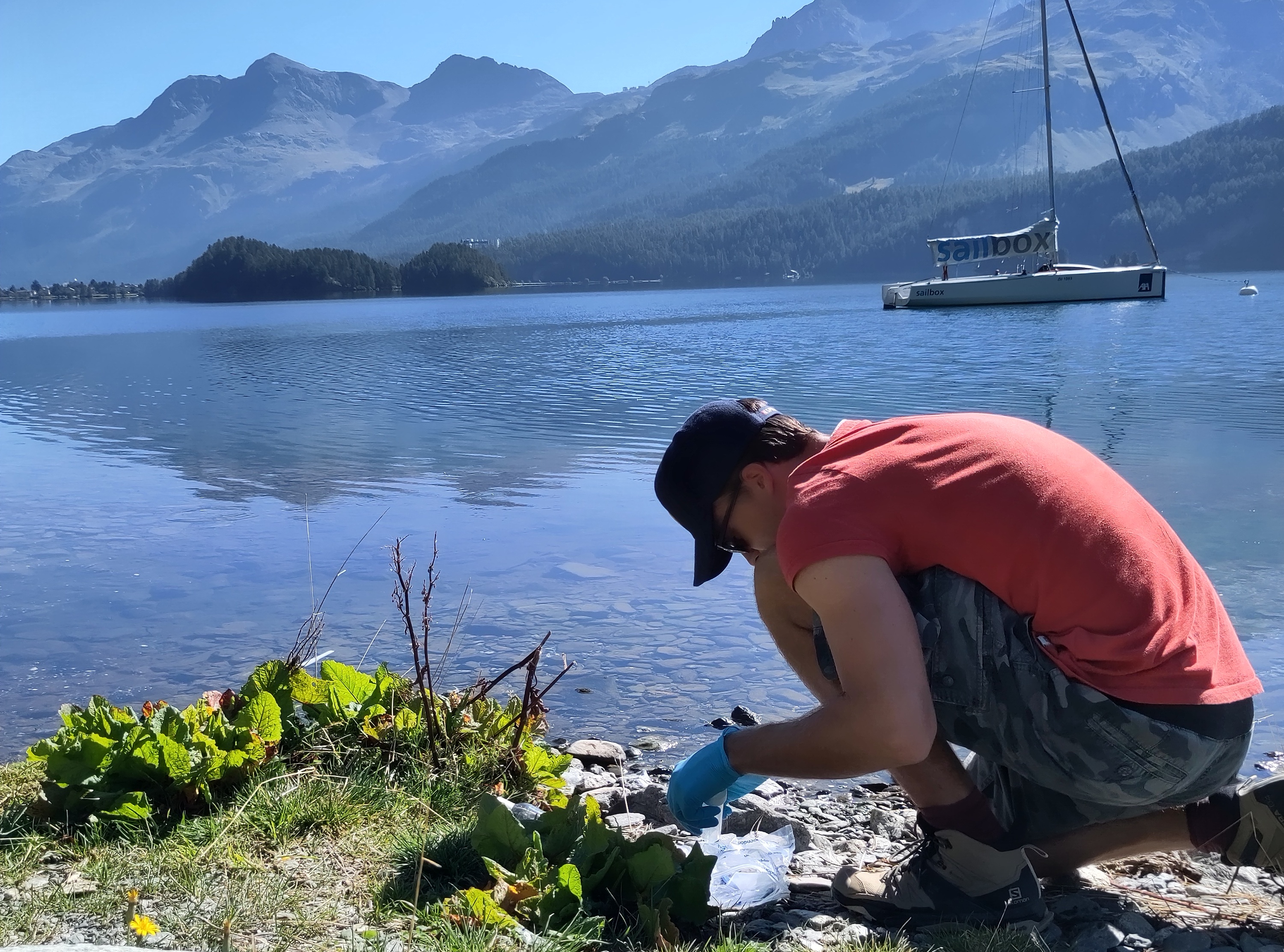 A man wearing gloves crouches by a lake, about to collect samples of the water,