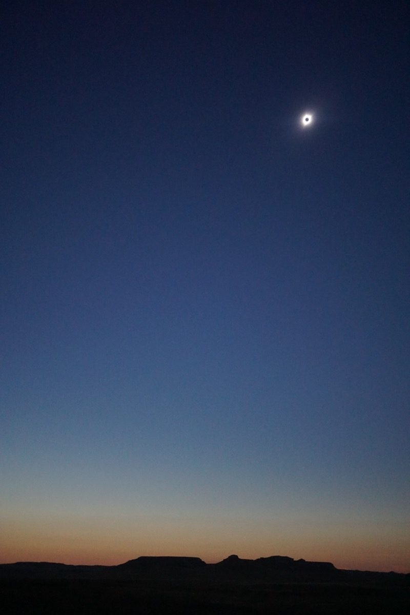 A total eclipse of the Sun is shown in a deep blue sky high above the silhouette of mountains.
