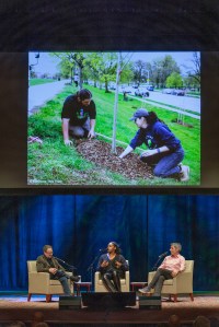 Three people on stage with a photo of two people planting a tree projected above them.