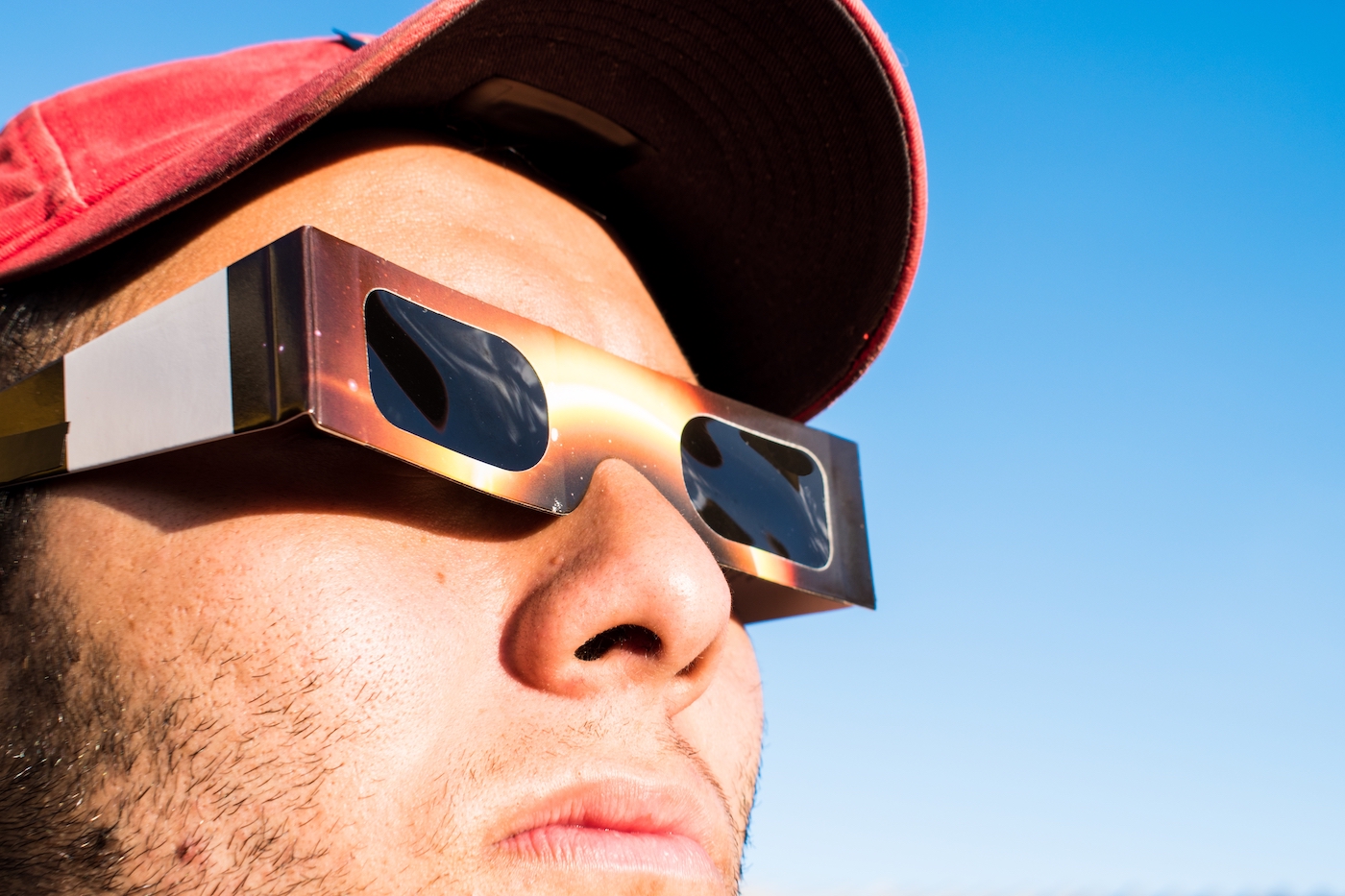 Portrait of man wearing cap and solar eclipse glasses, looking sun with blue sky at background