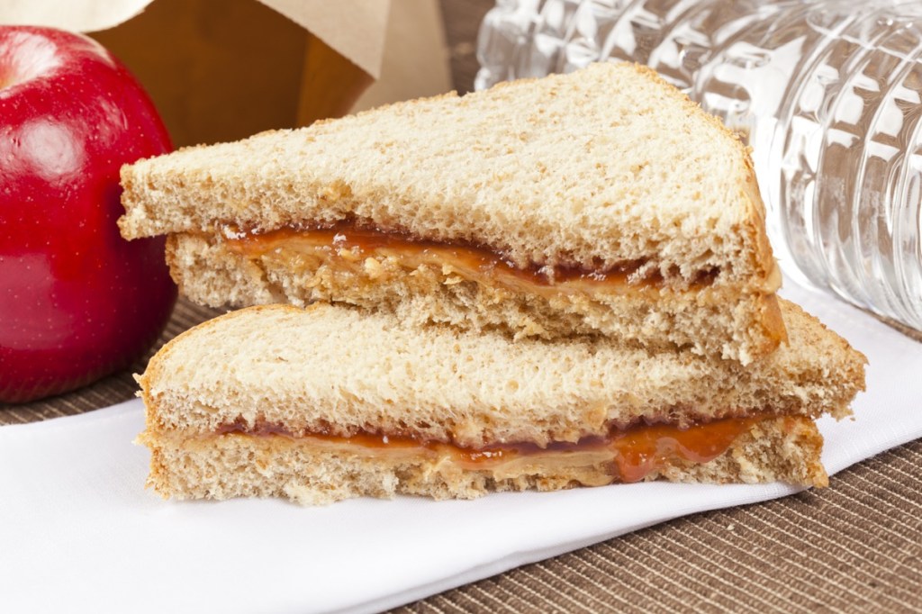 A close-up of a peanut butter and jelly sandwich on whole wheat bread, sitting on a napkin with an apple, and a water bottle nearby.