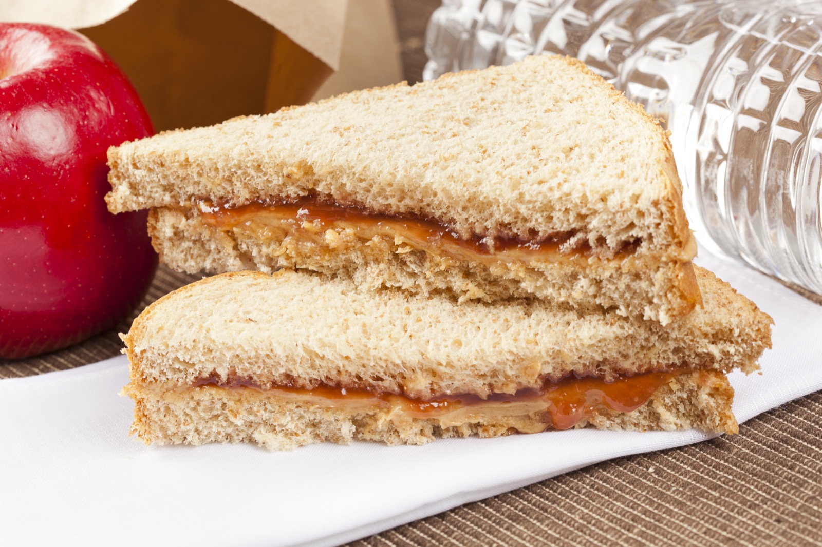 Un primer plano de un sándwich de mantequilla de maní y mermelada en pan integral sobre una servilleta, con una manzana y una botella de agua cerca.