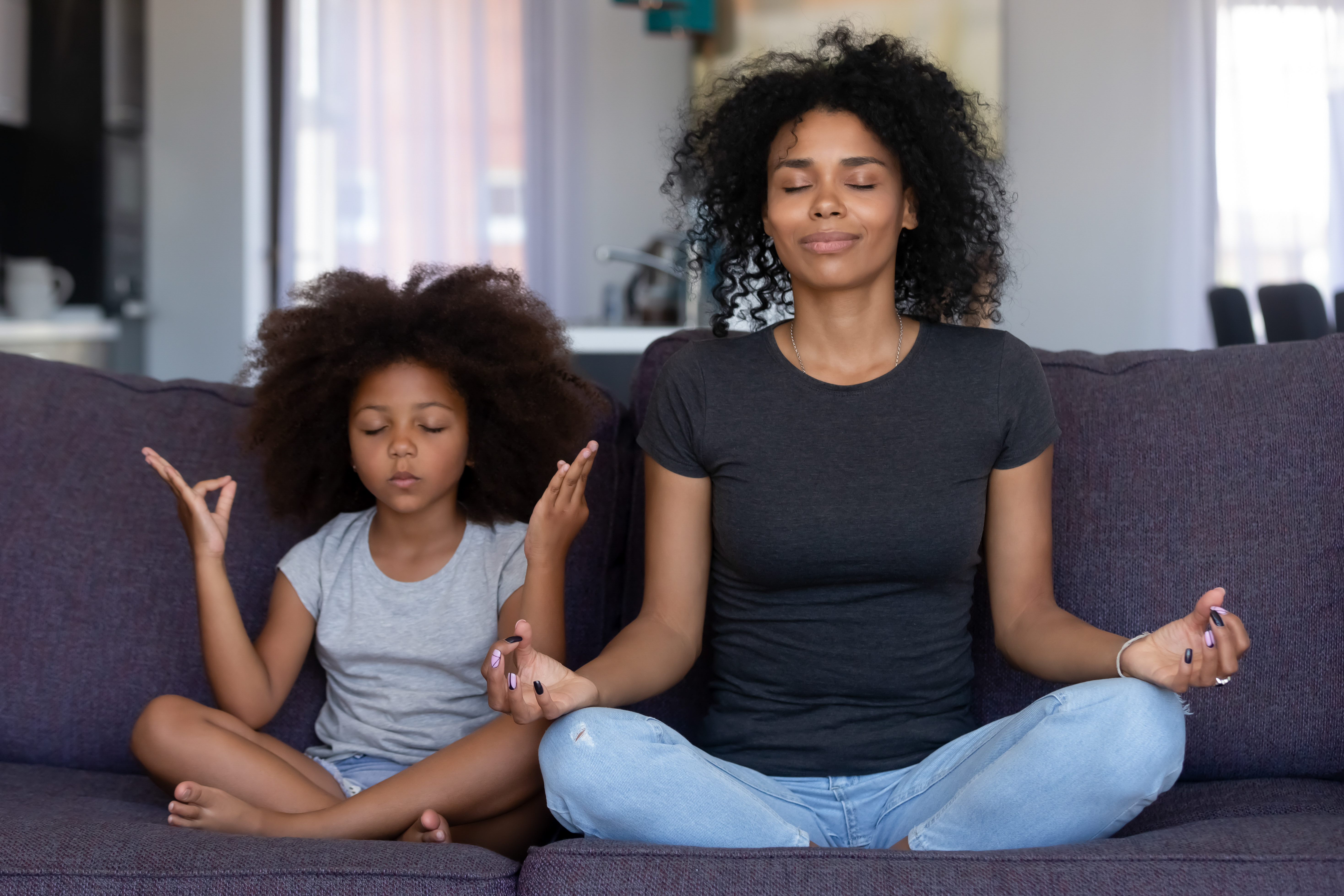 A parent and child sit on a couch with crossed legs, hands on their knees, and their eyes closed as they meditate